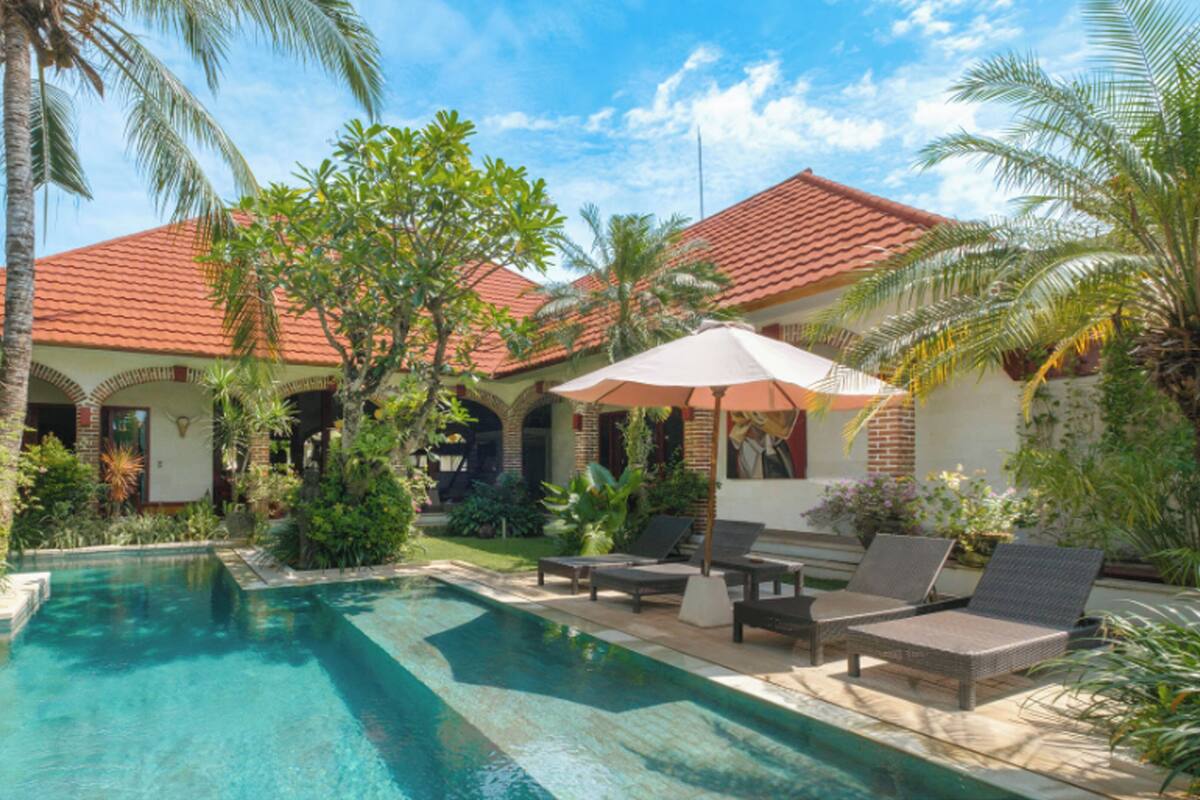 Área de piscina com espreguiçadeiras e um guarda-sol, rodeada por palmeiras e um edifício com telhado de telhas vermelhas.