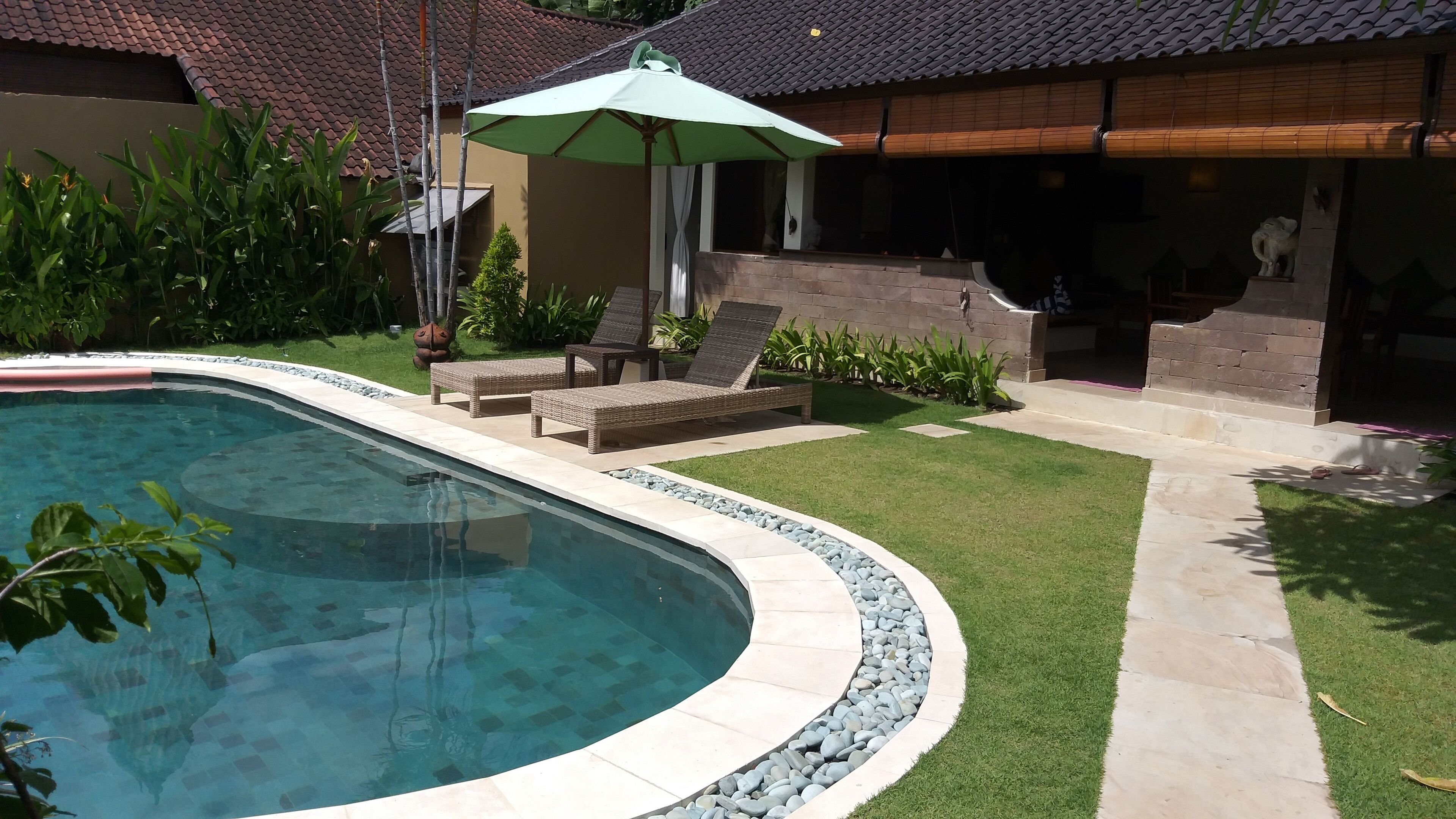 Une piscine entourée de chaises longues et d’un parasol, le tout bordé d’une pelouse bien entretenue et d’un mur de pierres.