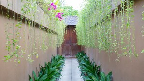 A narrow pathway flanked by lush green plants and hanging vines, leading to a traditional wooden door. - Dyana Villas (Seminyak)