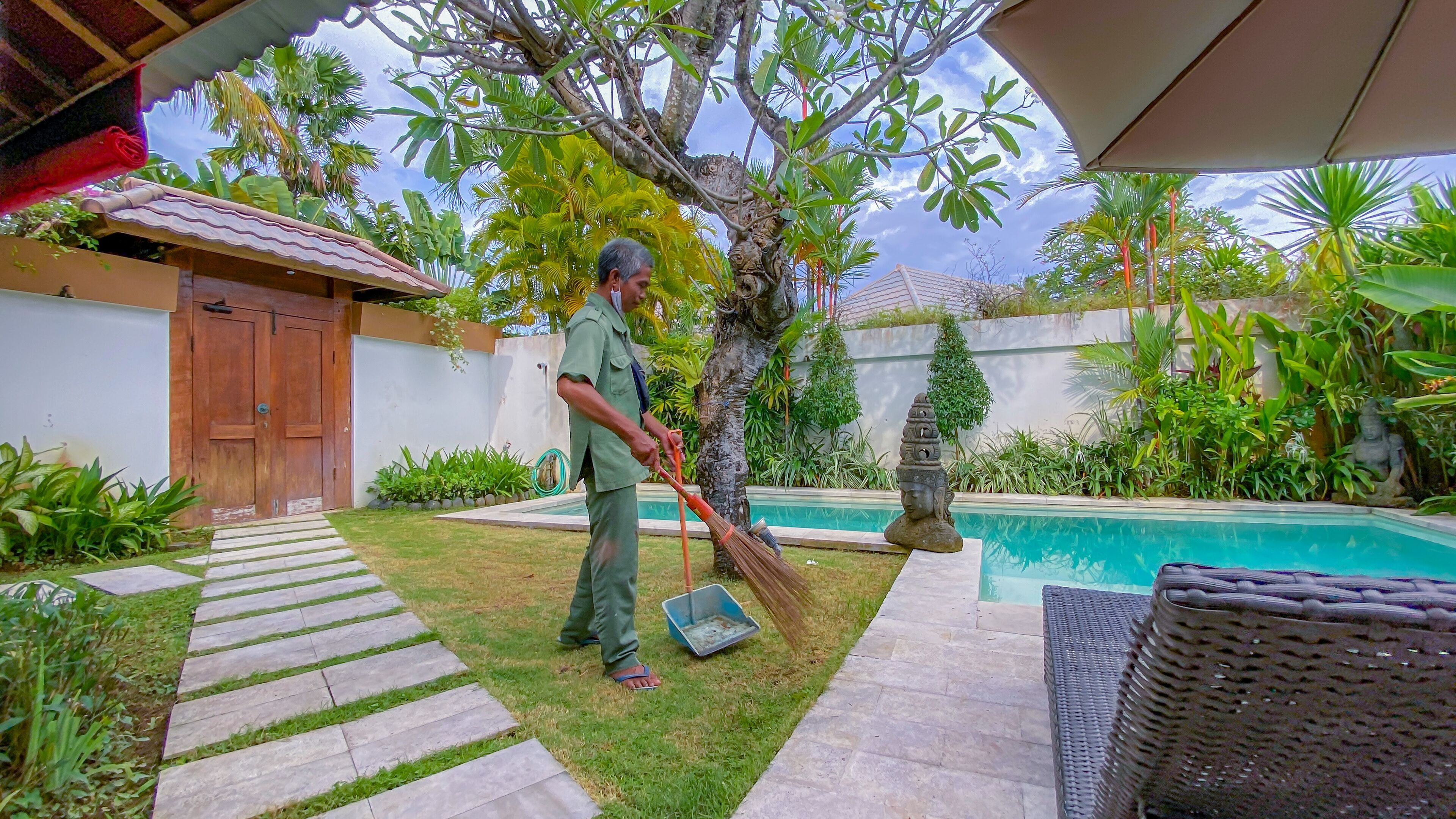 Un homme balaie une terrasse à l’aide d’un balai et d’une pelle. On aperçoit une piscine, une statue et diverses plantes en arrière-plan.