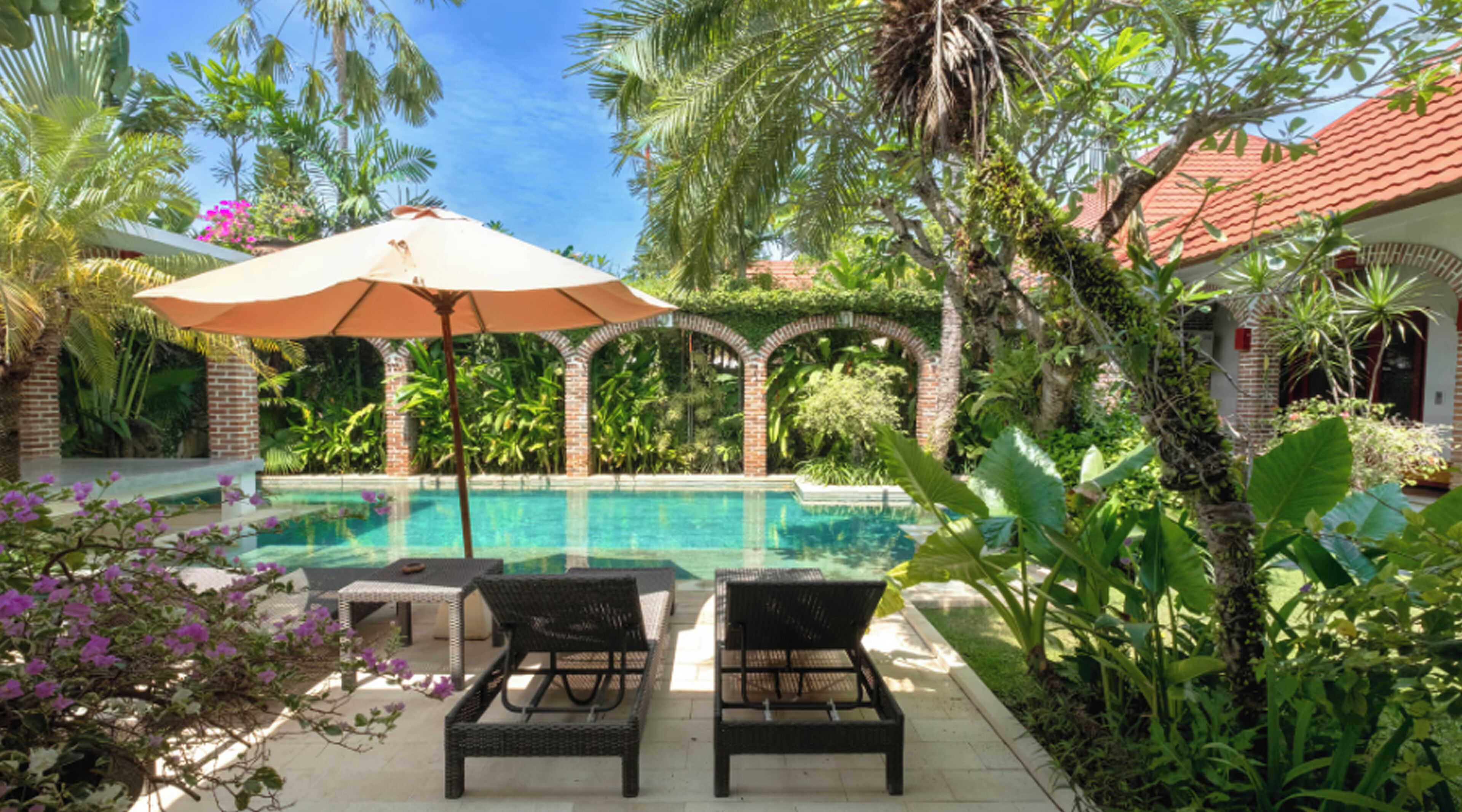 Un espace aménagé au bord de la piscine, avec deux chaises longues, un grand parasol et une végétation tropicale luxuriante.