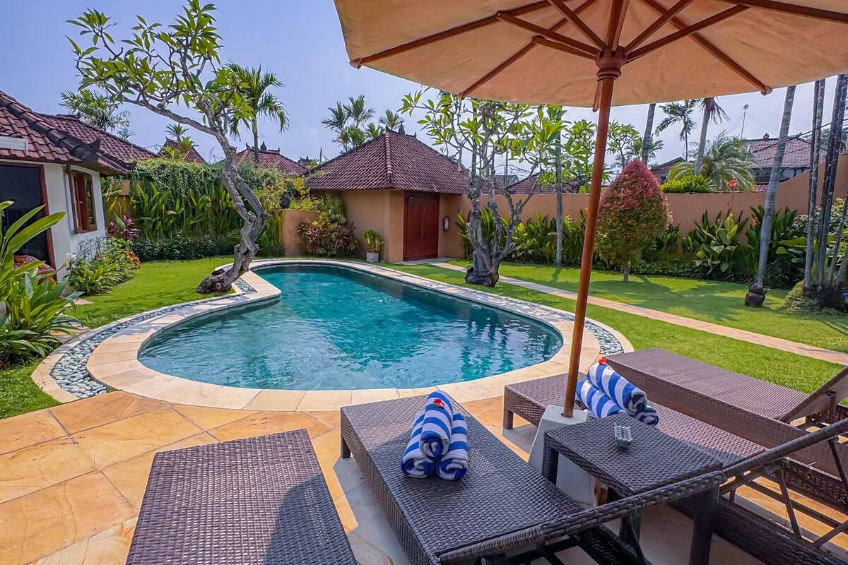 Piscina com espreguiçadeiras e um guarda-sol, rodeada por vegetação e um deck revestido com azulejos.