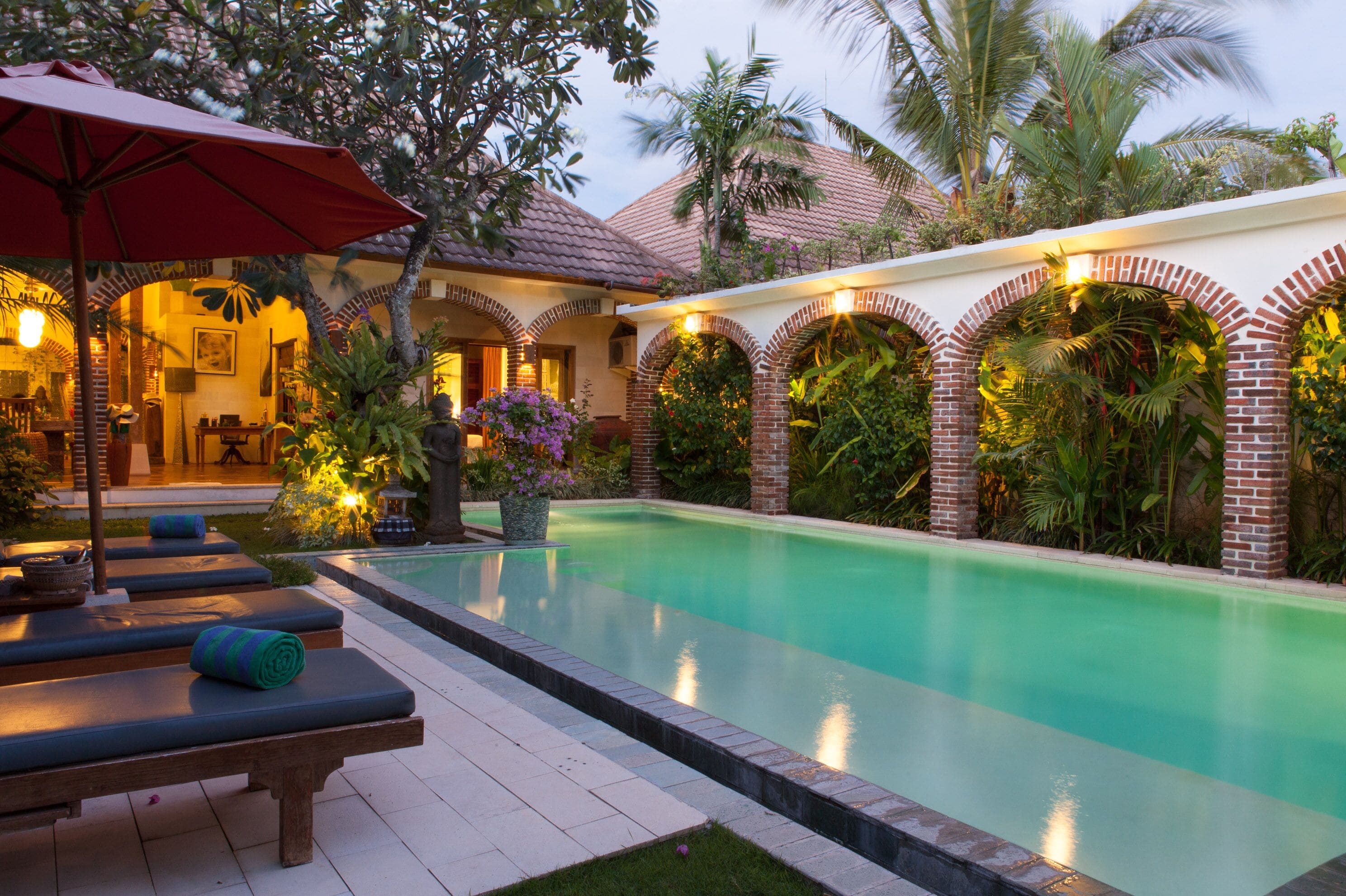 Un espace aménagé au bord de la piscine, avec des chaises longues et un parasol, entouré d’une végétation luxuriante et d’un bâtiment en briques doté d’arcades.