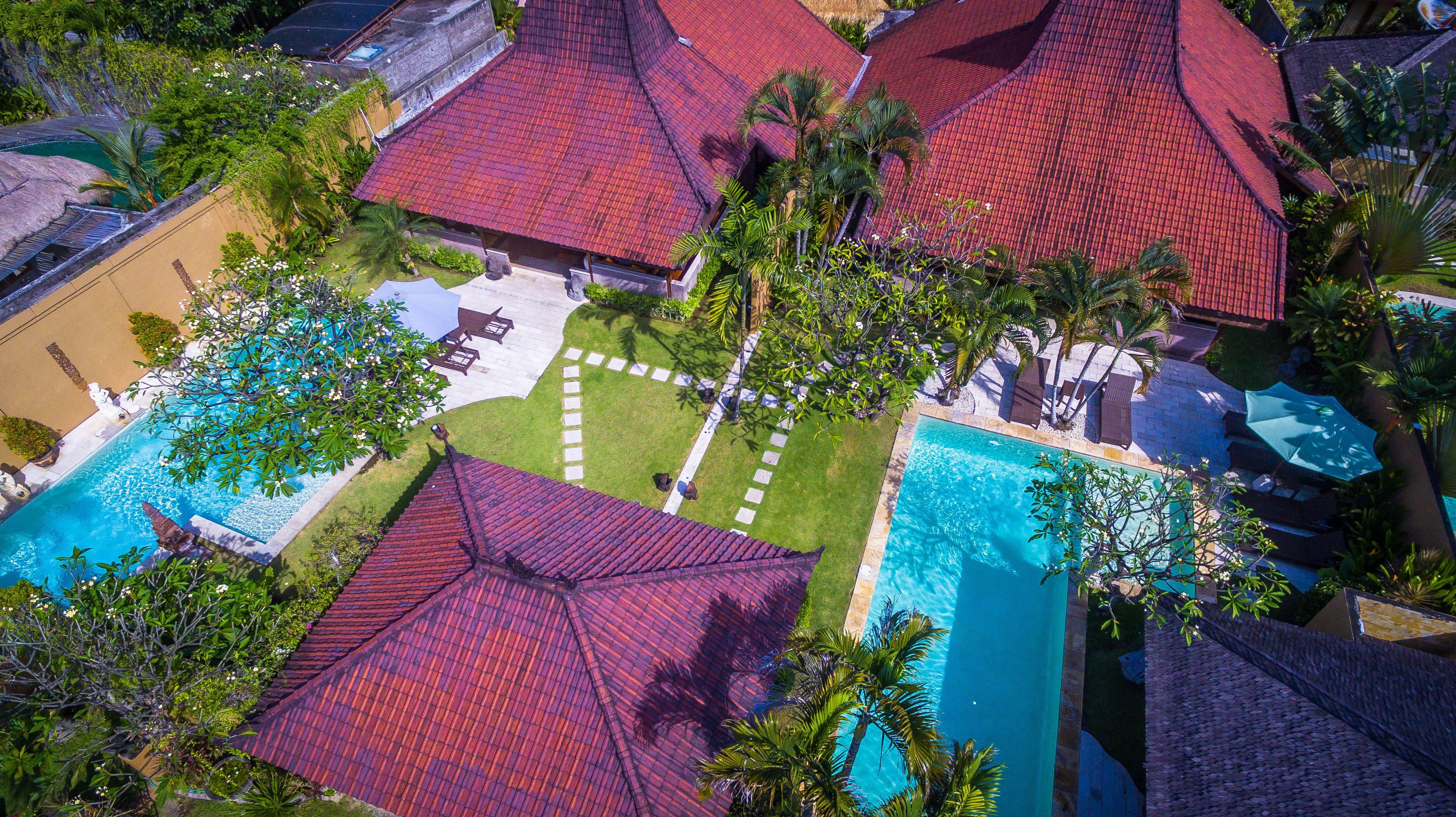 An aerial view of a resort with multiple red-roofed buildings, a central swimming pool, and surrounding greenery.