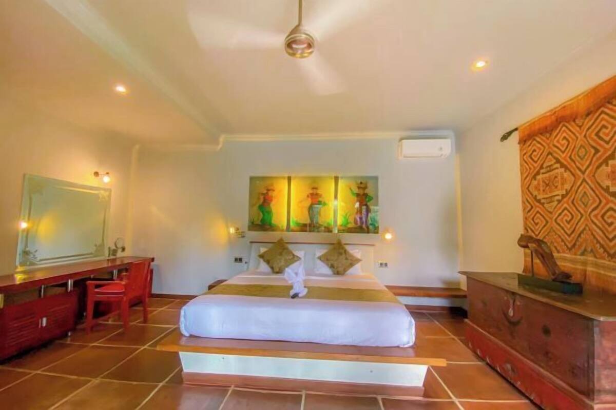 Quarto de hotel com cama, escrivaninha, cadeira, espelho e um objeto decorativo na parede.