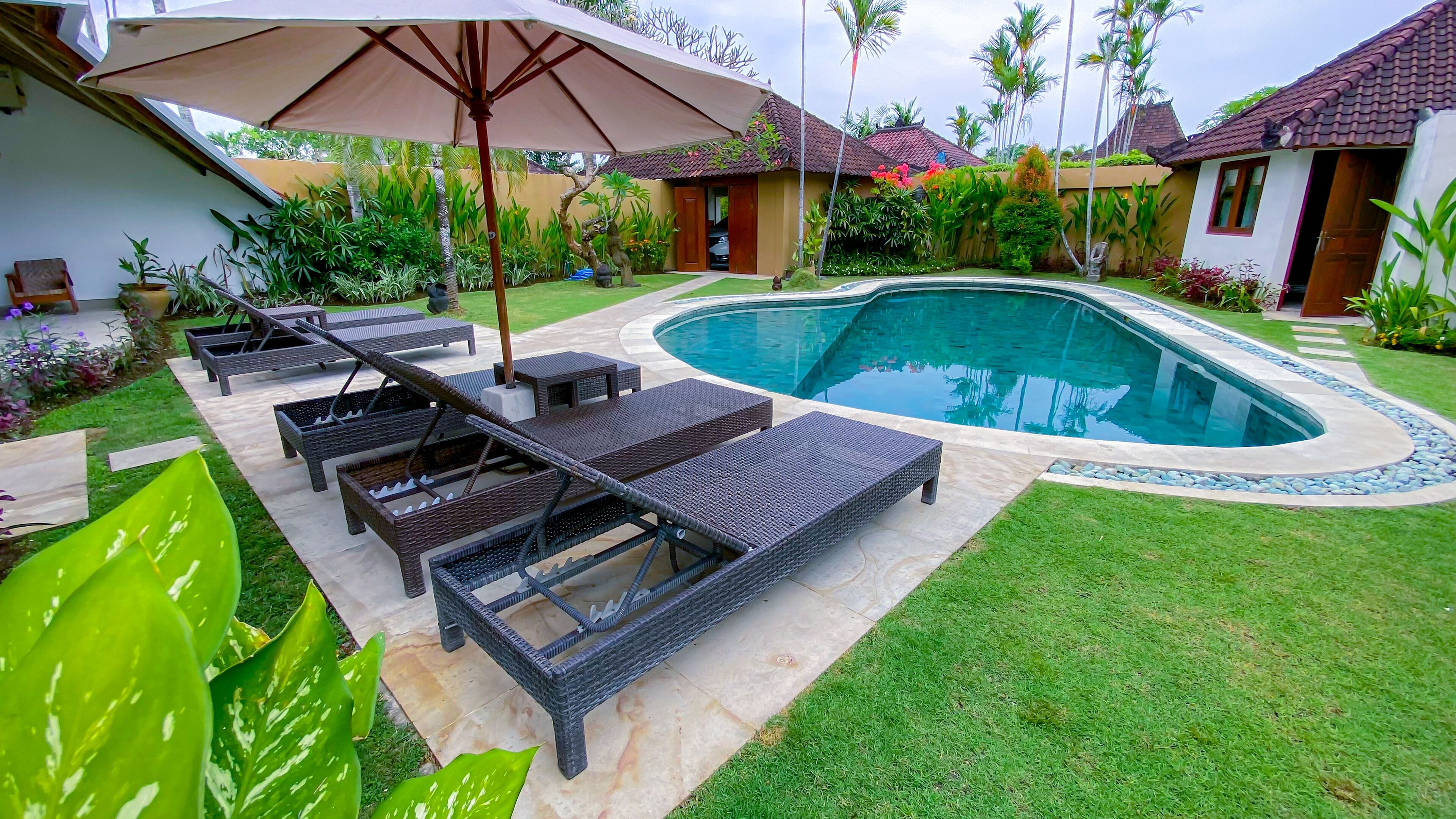 A poolside area with lounge chairs and an umbrella, surrounded by well-maintained greenery and a swimming pool.