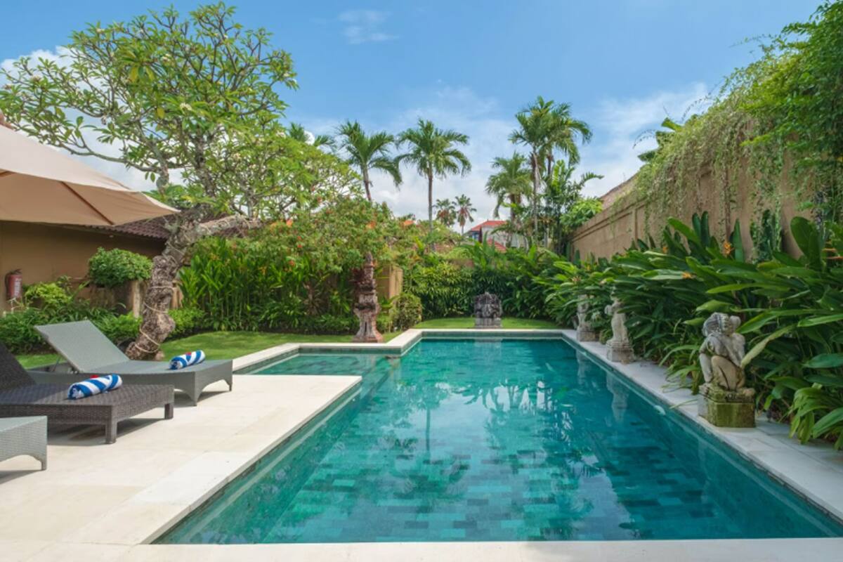 Uma piscina retangular com água azul cristalina, rodeada por vegetação exuberante, incluindo palmeiras e diversas plantas. Há espreguiçadeiras e uma pequena estátua perto da piscina.