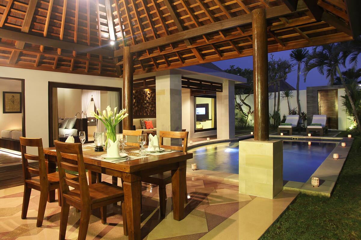 An outdoor dining area with a pool, wooden furniture, and a covered seating area.