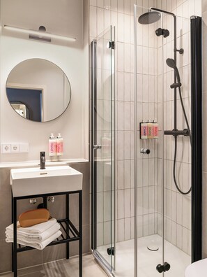 A modern bathroom with a glass-enclosed shower, a round mirror, and a sink with toiletries. - Numa Munich St Paul (Munich)