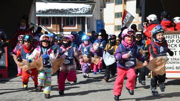 A group of children in winter sports attire walking in a line, carrying bags, with a sign reading "SHUTTLE TOP SCHOOL" in the background.