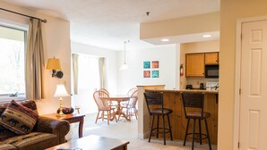 A living room with a sofa, coffee table, and a dining area with chairs and a table. There is a kitchen area with a bar and stools. - Vantage Point Villas at Stratton Mountain Resort (Stratton)