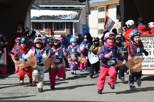 Un grupo de niños con ropa de deportes de invierno caminando en fila, llevando bolsas, con un cartel que dice "SHUTTLE TOP SCHOOL" al fondo.