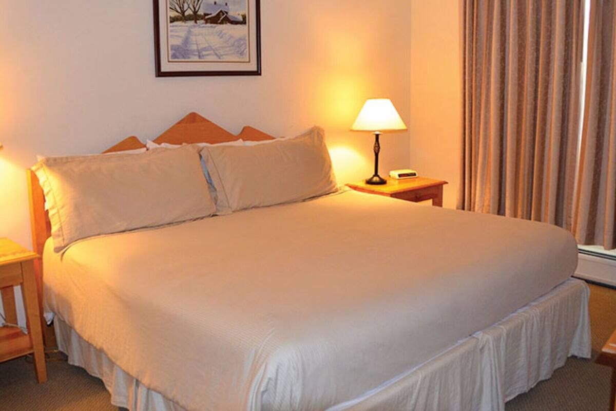 A neatly made bed with a headboard, two bedside lamps, a framed picture on the wall, and a window with curtains.