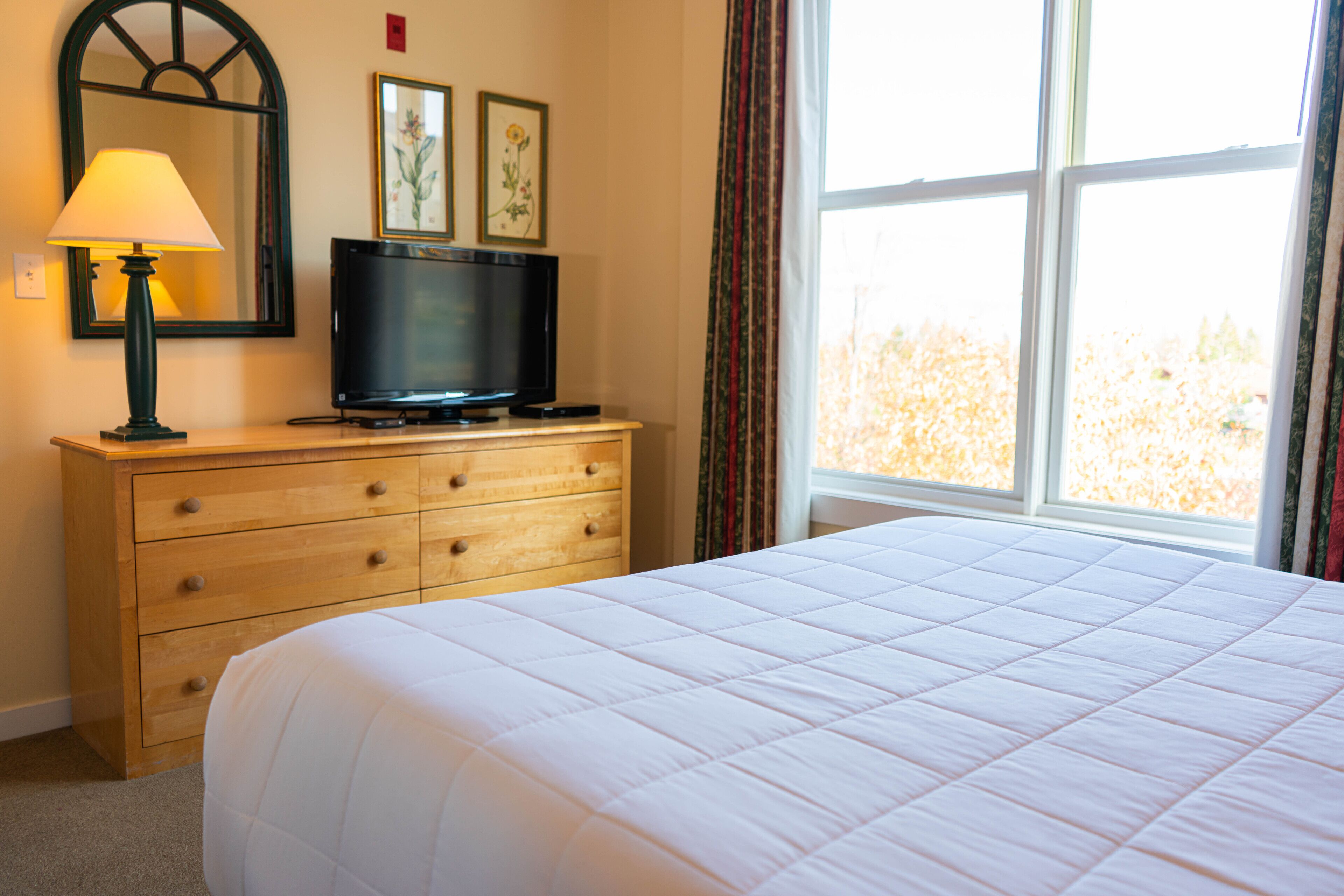 A hotel room with a bed, a wooden dresser, a television, and a window with curtains.