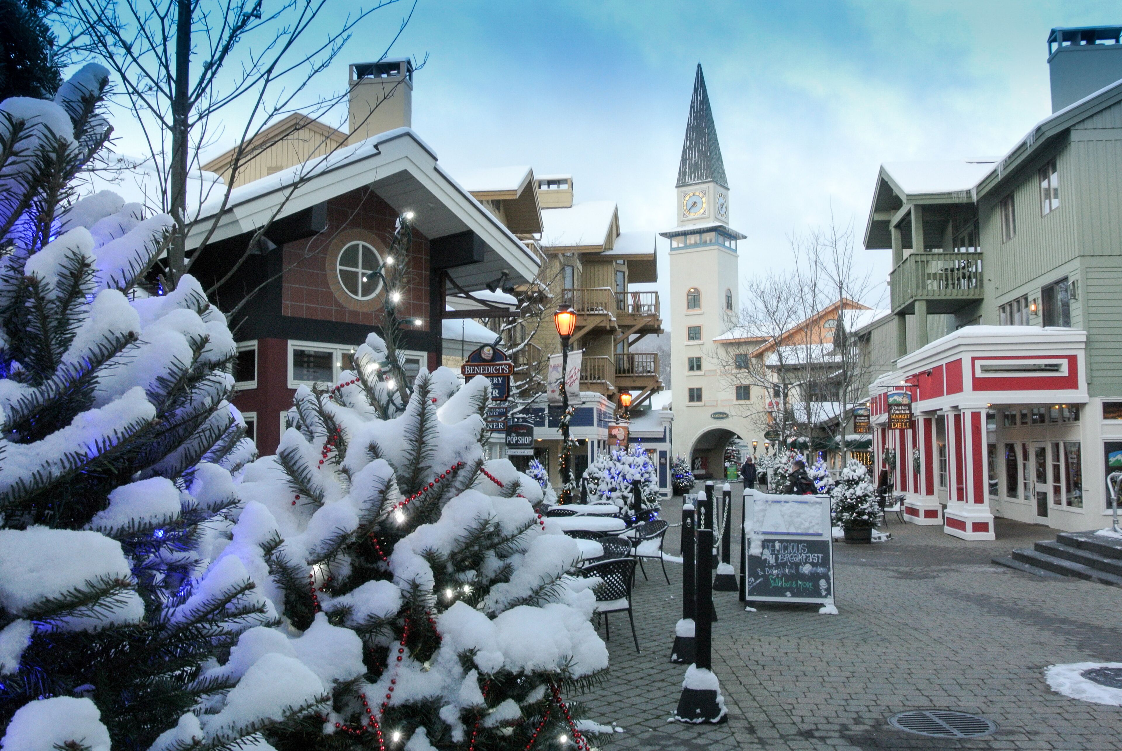 Ein verschneiter Stadtplatz mit Gebäuden, einem Glockenturm und Weihnachtsbäumen.