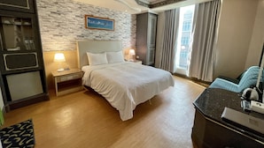 Une chambre d’hôtel comprenant un lit, des tables de chevet, un bureau, une chaise, une fenêtre avec des rideaux et un mur en pierre orné d’un tableau.