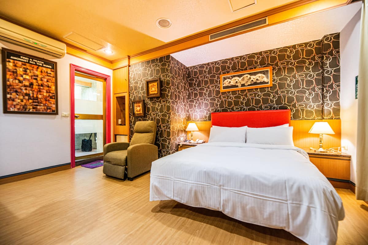 Quarto de hotel com cama grande, uma cadeira, escrivaninha com abajur e parede com padrão.