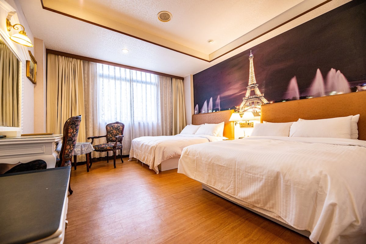 Quarto de hotel com duas camas, uma escrivaninha, uma cadeira e uma pintura grande da Torre Eiffel.