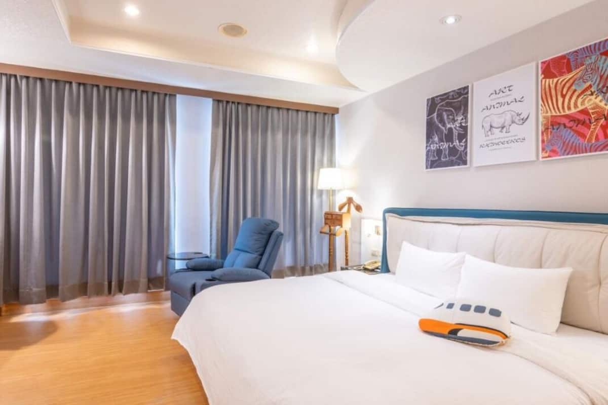 Quarto de hotel com uma cama grande, uma cadeira, uma escrivaninha e cortinas.
