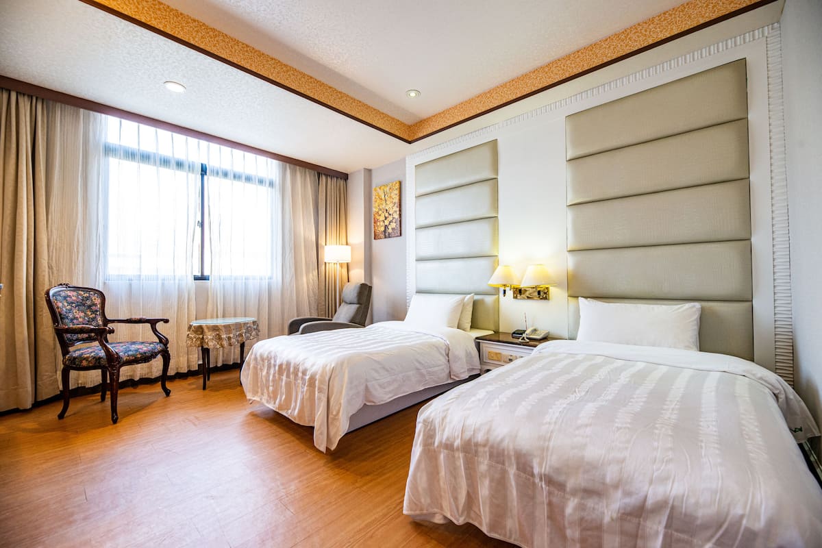 Quarto de hotel com duas camas, uma cadeira, uma mesinha e um abajur.