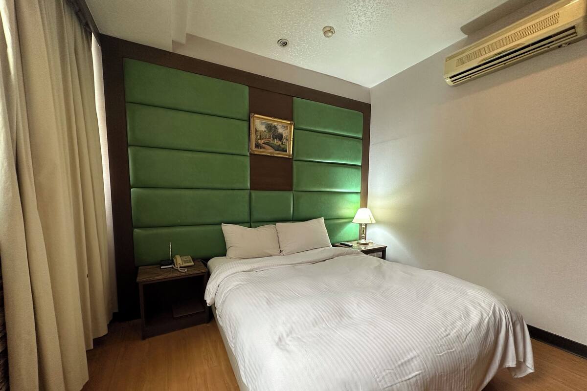 Quarto de hotel com uma cama grande, cabeceira acolchoada verde, luminária de cabeceira, mesa de cabeceira e um quadro emoldurado na cabeceira.
