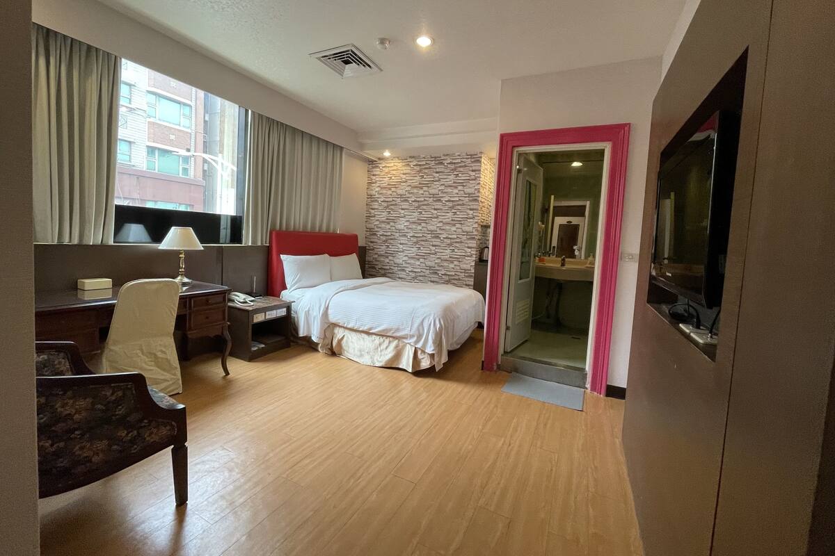 Quarto de hotel com cama, escrivaninha, cadeira, televisão e banheiro visível através de uma porta aberta.