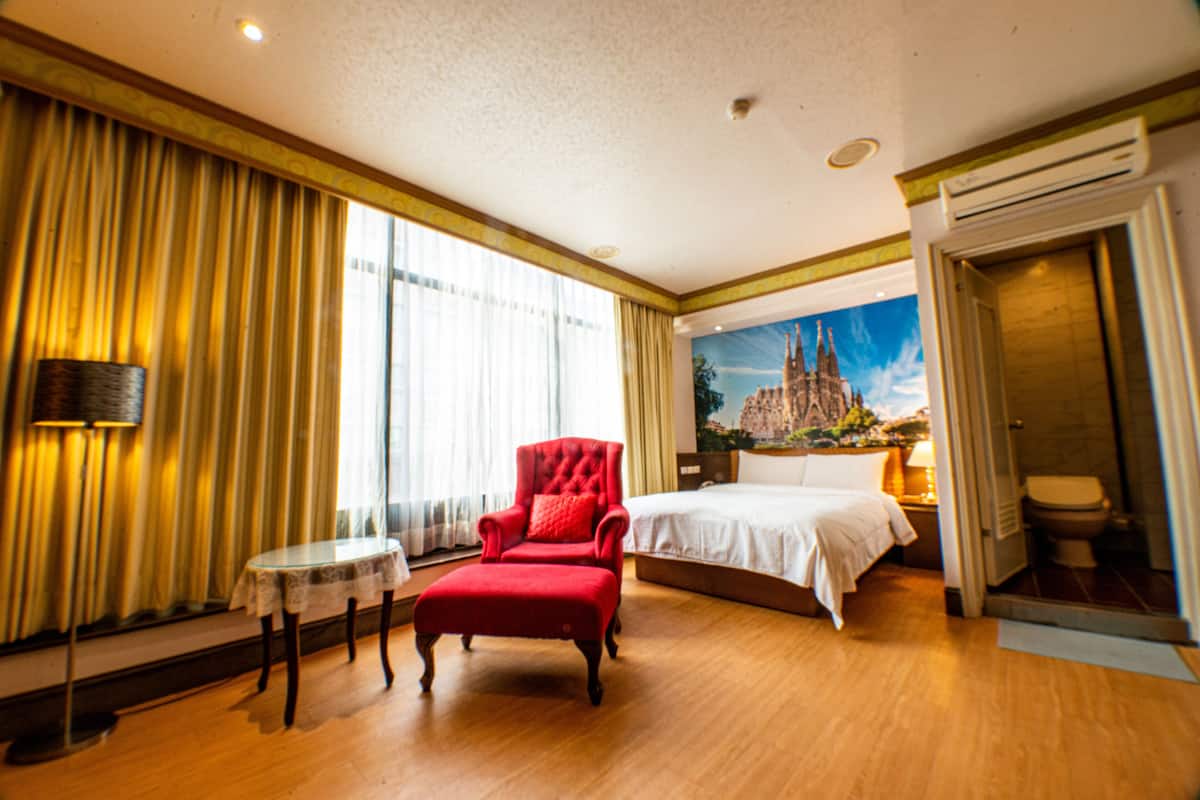 Quarto de hotel com uma cama grande, uma poltrona vermelha, uma mesa pequena e um quadro de um castelo na parede.