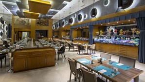 A spacious restaurant with multiple dining tables and chairs, a central bar area with a variety of food displays, and a modern ceiling design with recessed lighting.