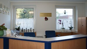 Zona de recepción de un hotel con un mostrador de madera, dos sillas azules, un reloj y un cartel dando la bienvenida a los huéspedes.