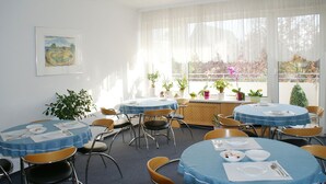 Comedor con mesas redondas, sillas y manteles azules. Hay plantas en macetas y una ventana con vista a árboles.