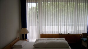 Una habitación de hotel con cama, mesita de noche, lámpara y ventana con cortinas translúcidas.