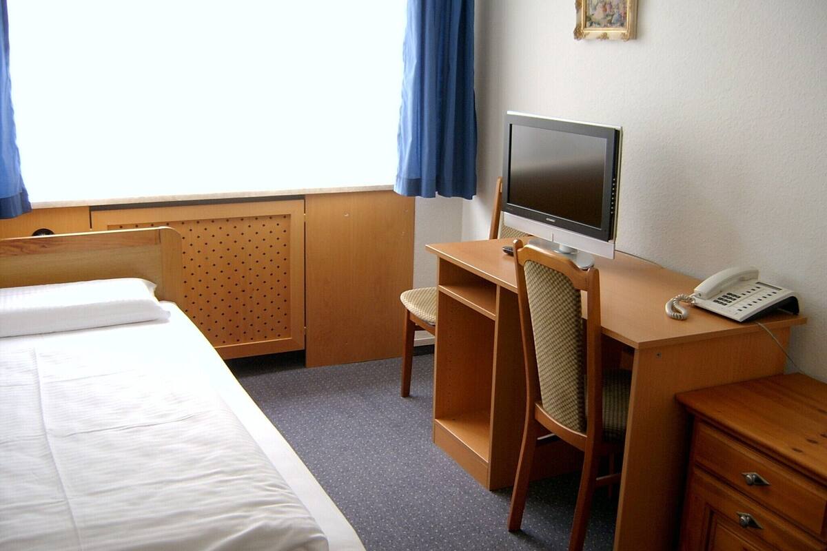 Hotellihuone, jossa on sänky, työpöytä tietokoneella, tuoli, puhelin ja taulu seinällä.