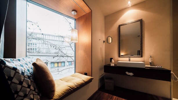 Ein modernes Badezimmer mit Holzboden, einem Fenster mit Blick auf Gebäude, einem Waschbecken mit Spiegel und einem Nachttisch mit Kissen.