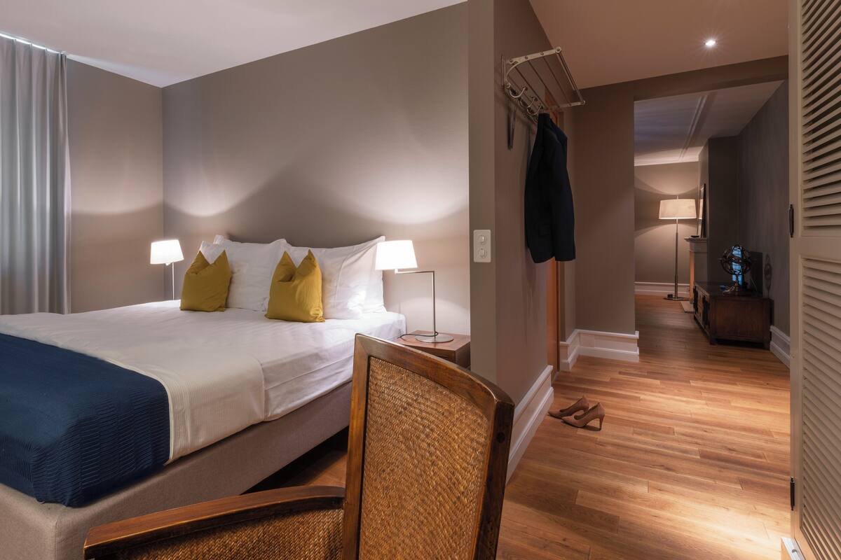 A hotel room with a bed, two bedside lamps, a chair, a coat rack, and a hallway leading to another room.