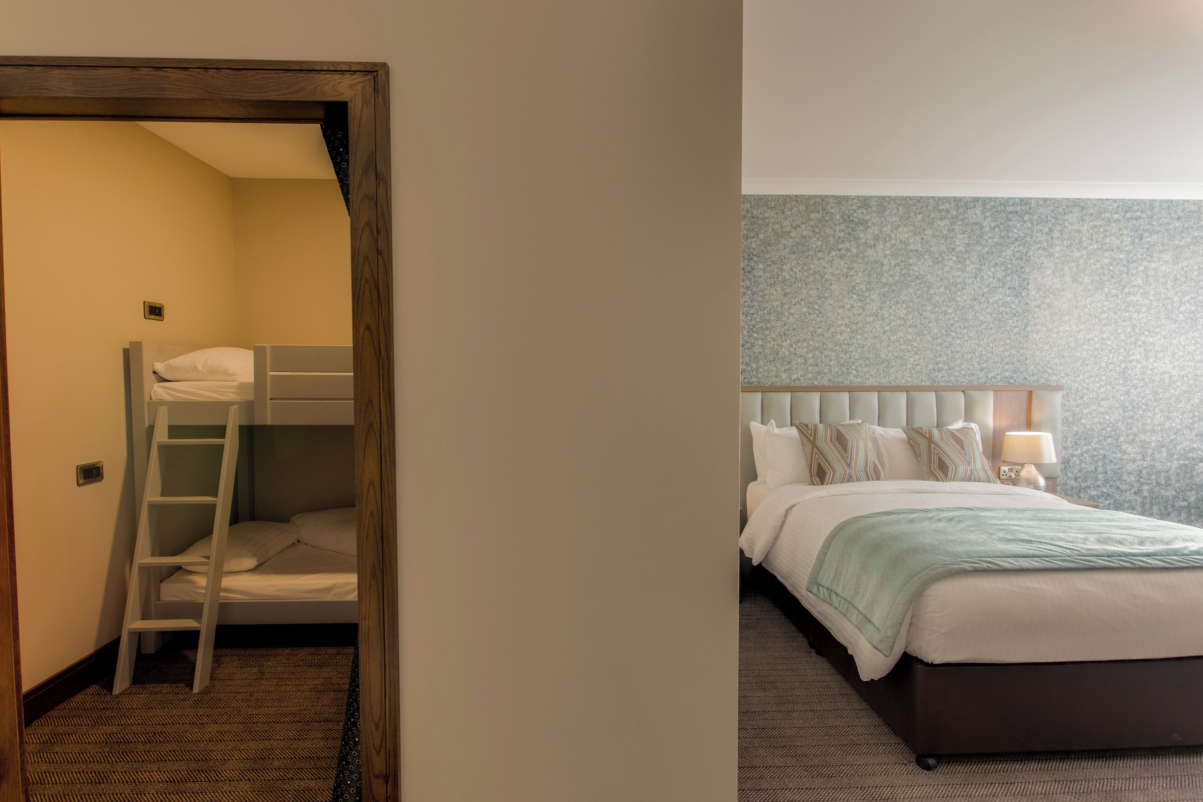 A room with a bunk bed, a ladder, a bedside table with a lamp, and a patterned wall.