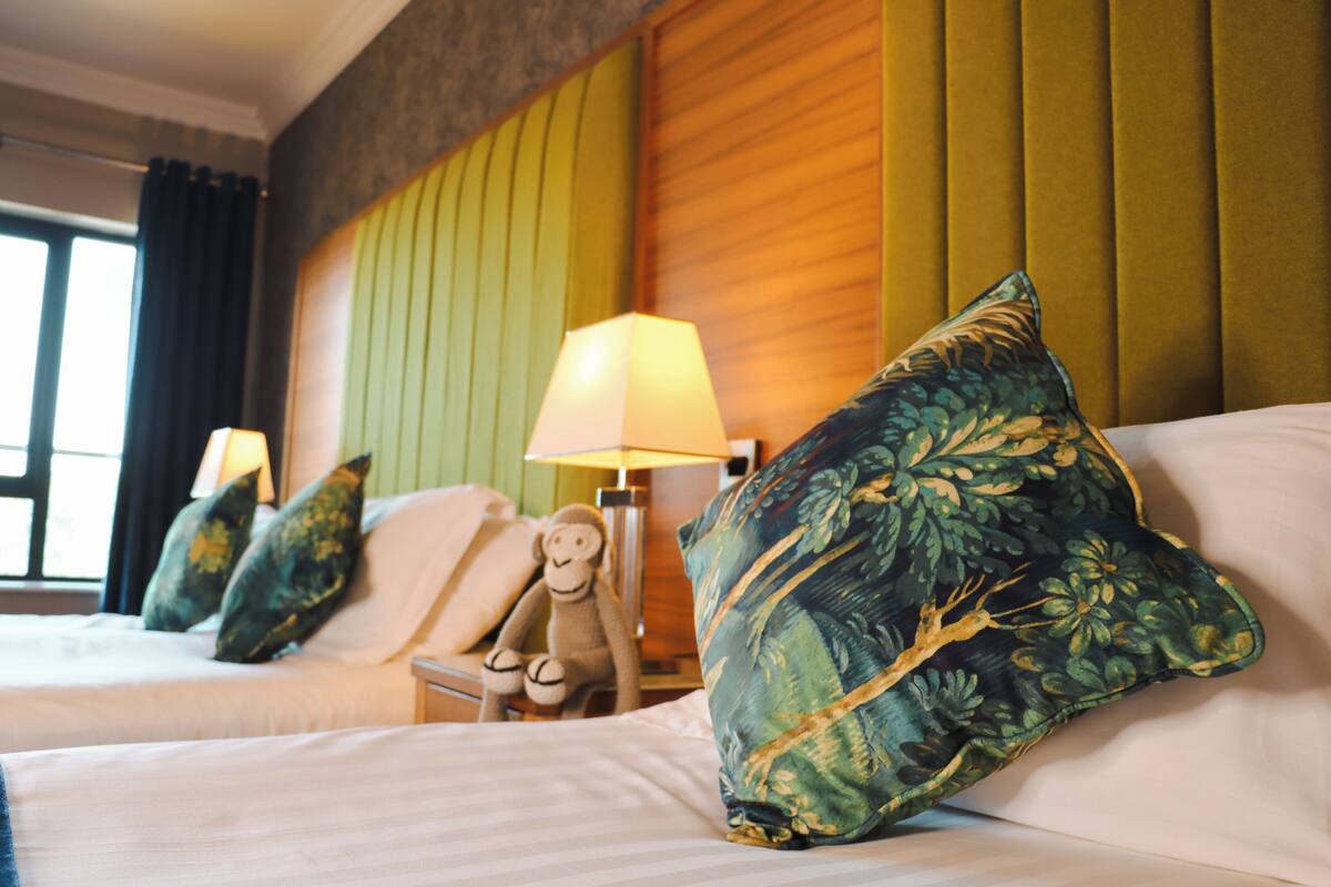 Ein Hotelzimmer mit zwei Betten, einem Nachttisch mit Lampe, einem weichen Affen-Plüschtier und einem großen, grünen Kissen.