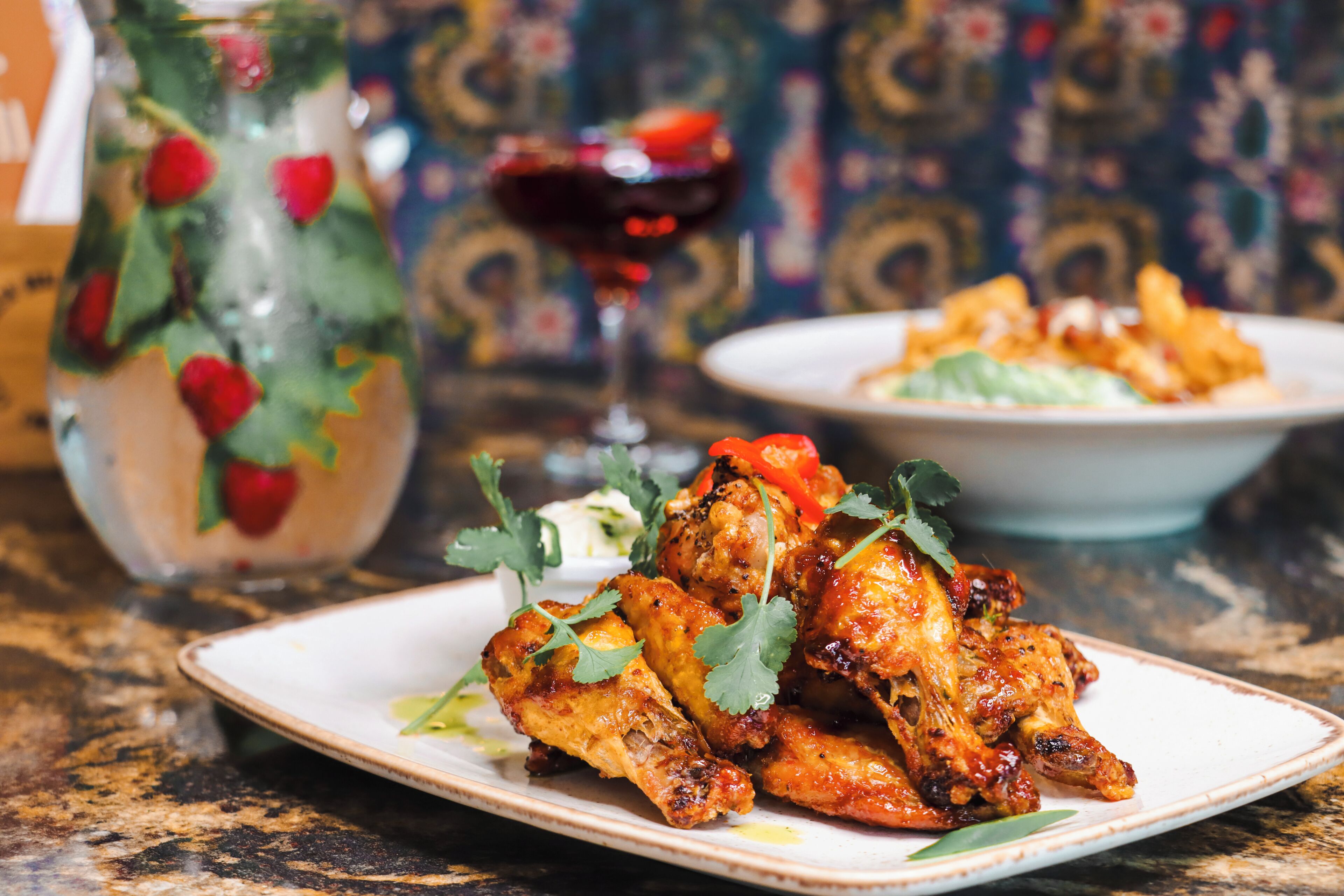 A plate of grilled chicken wings garnished with cilantro, a glass of red wine, and a pitcher with a strawberry garnish.