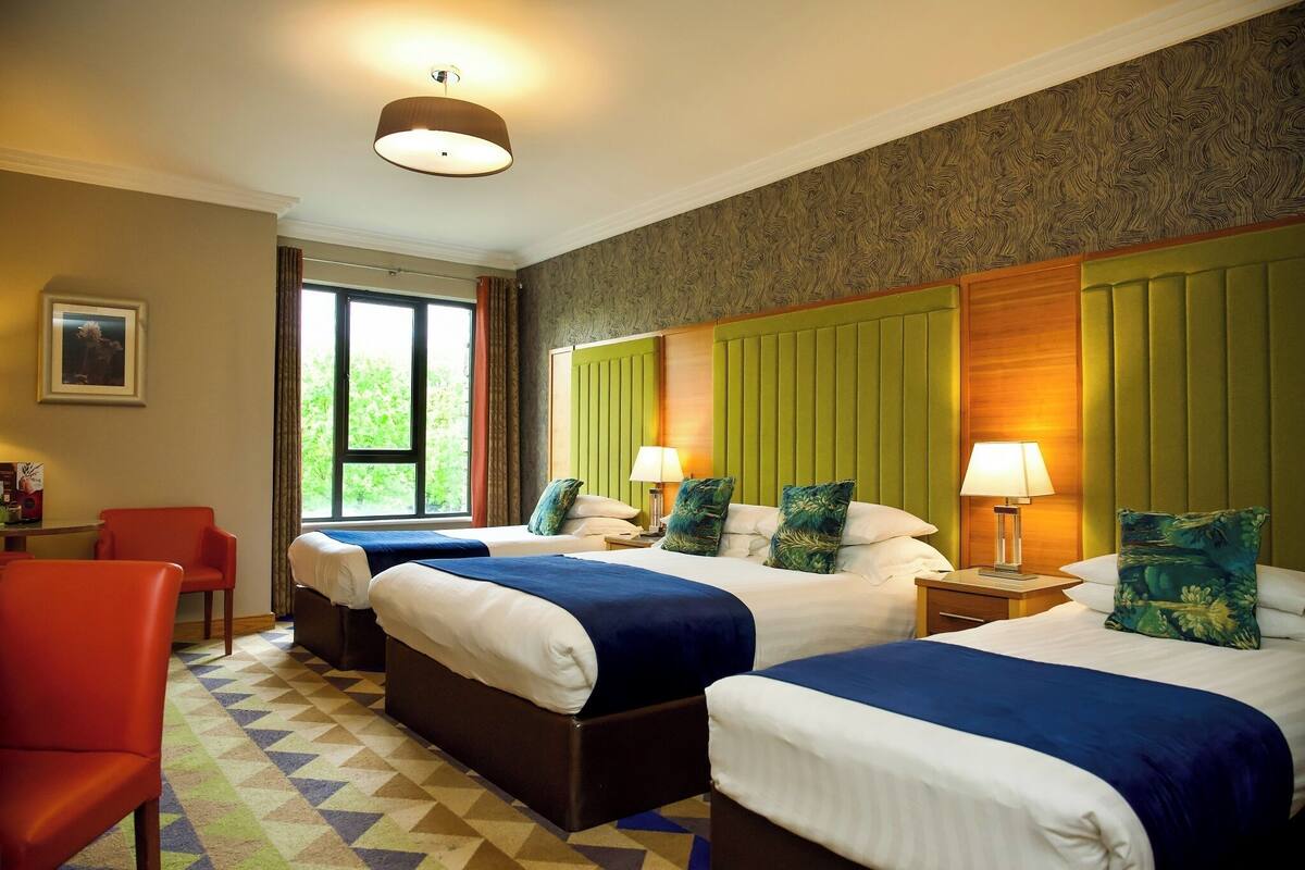 Ein Hotelzimmer mit zwei Betten, einem roten Sessel, einem Fenster mit Vorhängen und einer Wand mit Muster.