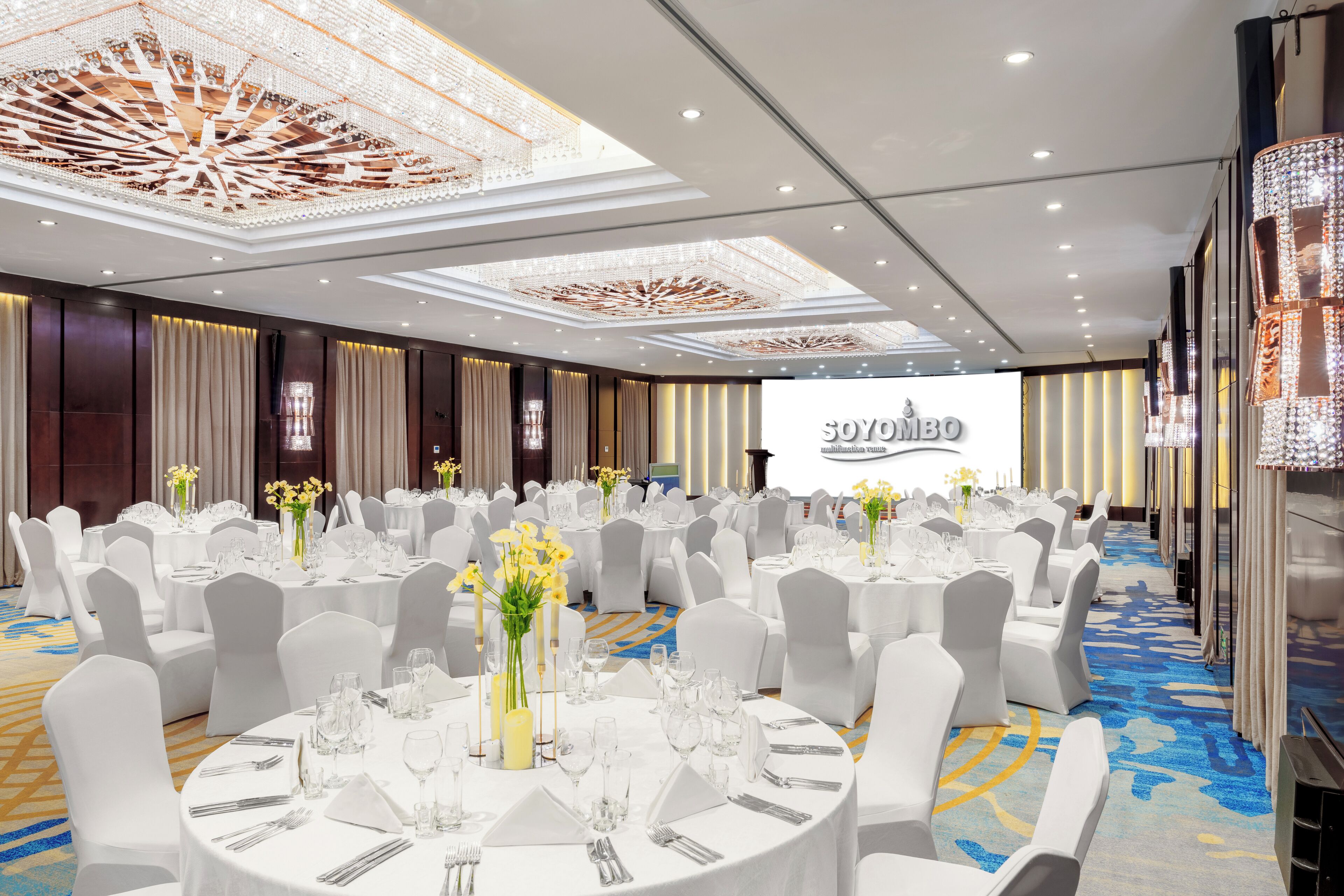 Un salón de banquetes con mesas redondas dispuestas para eventos, sillas blancas y candelabros en el techo.