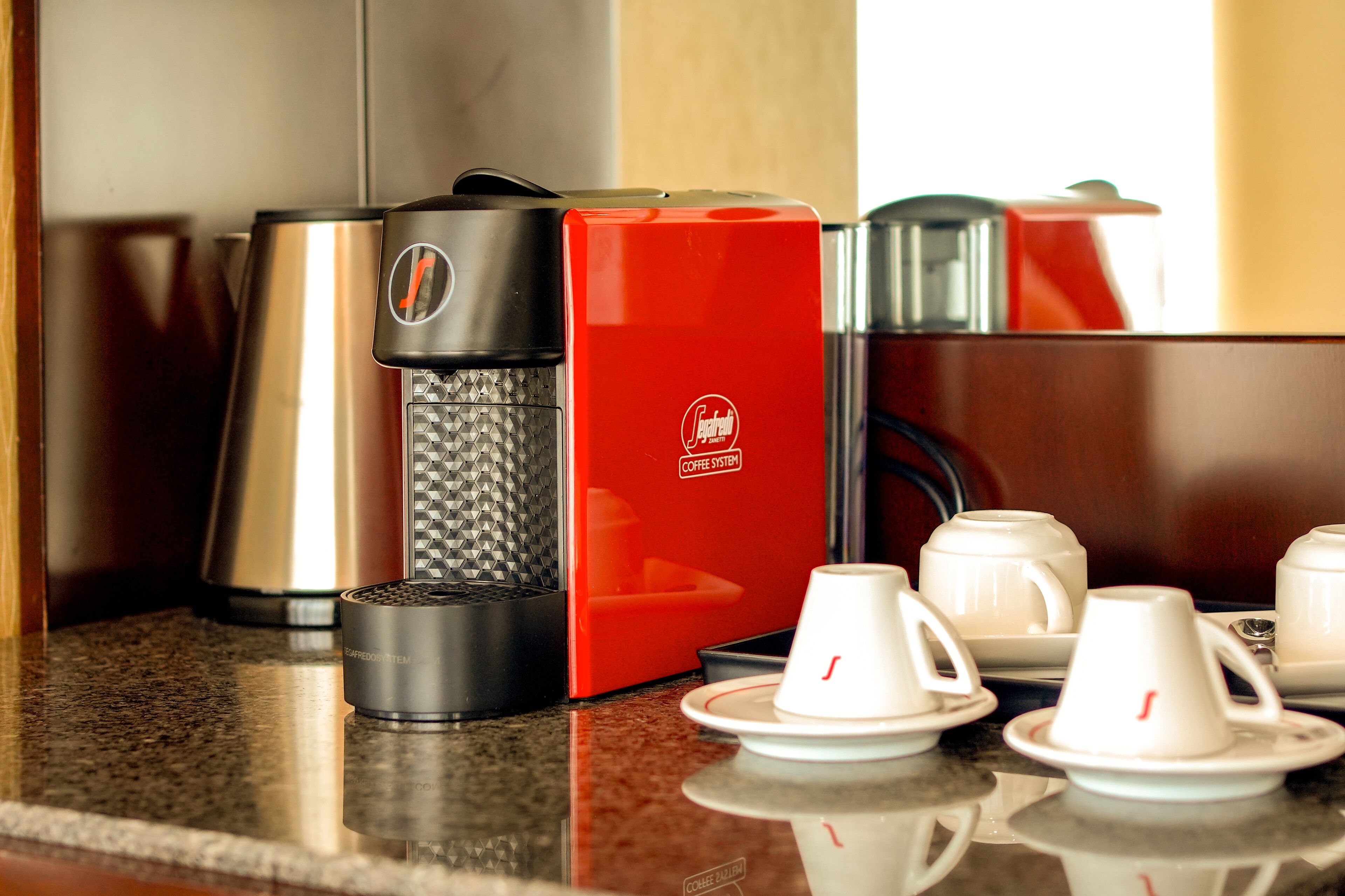 Una cafetera, un sistema rojo para preparar café y varias tazas blancas sobre una encimera.