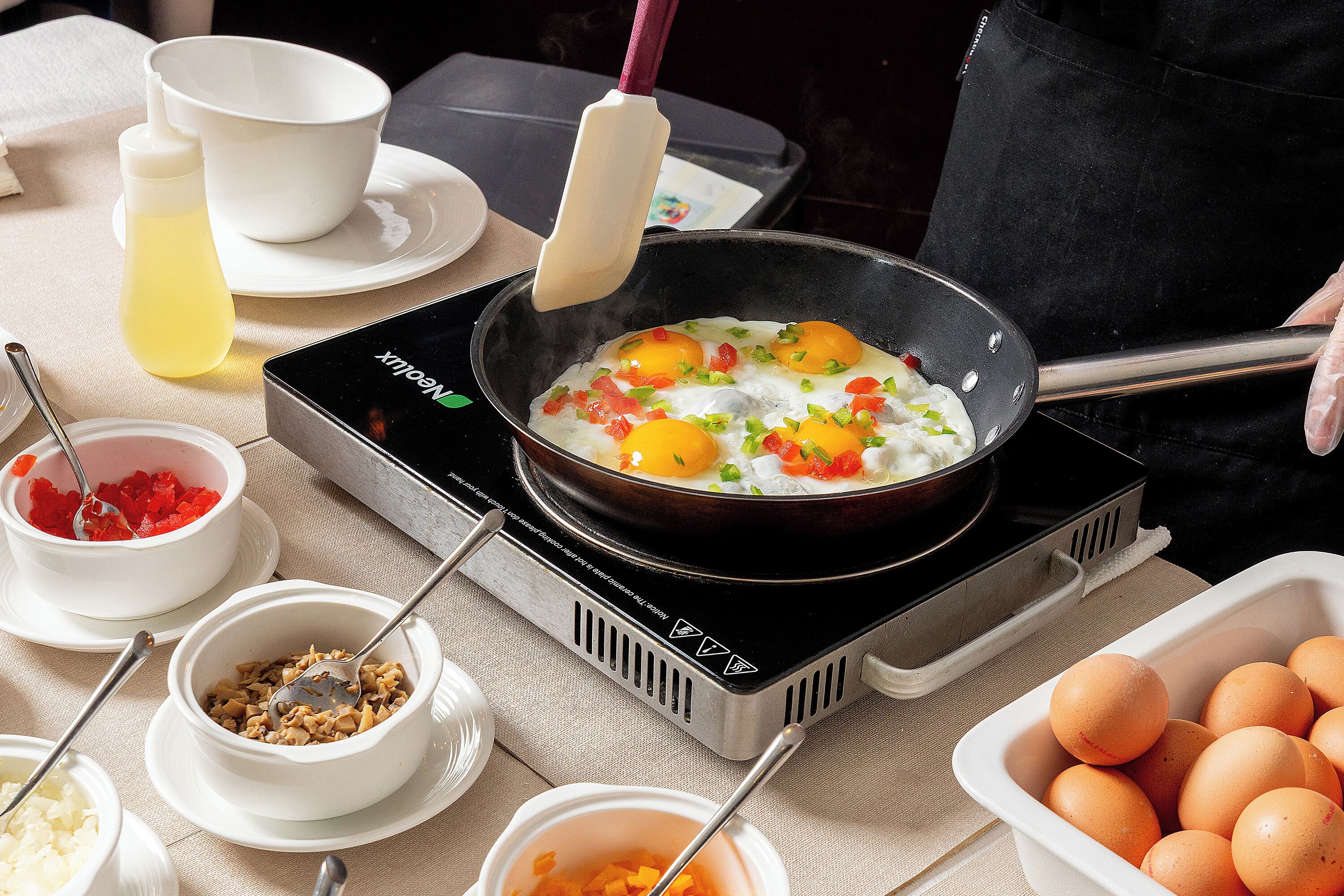 A kitchen with a frying pan on an induction cooktop, containing eggs and vegetables. There are bowls of ingredients and a container of eggs nearby.