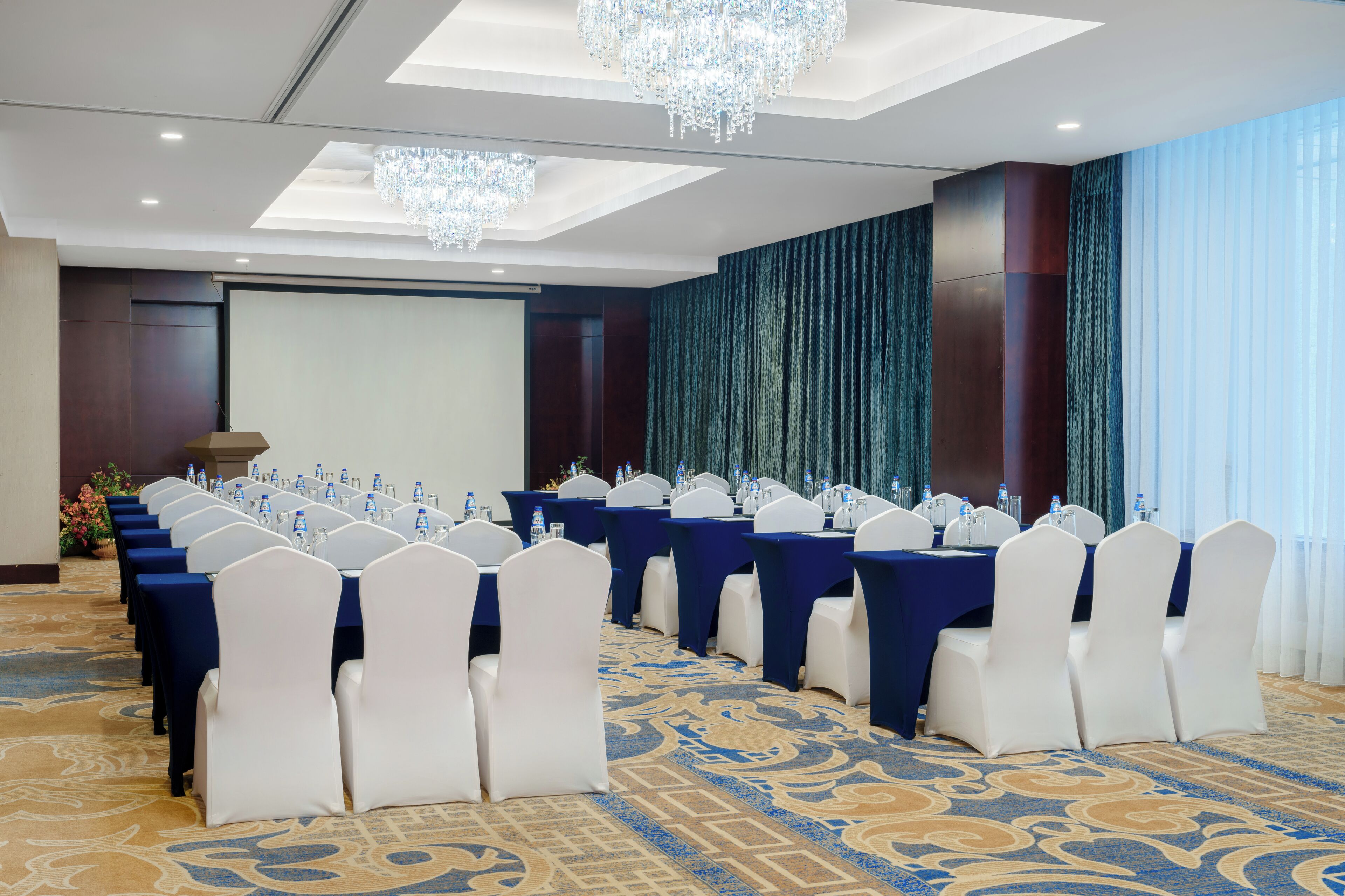 Un salón de conferencias con filas de mesas, sillas blancas y una pantalla grande.