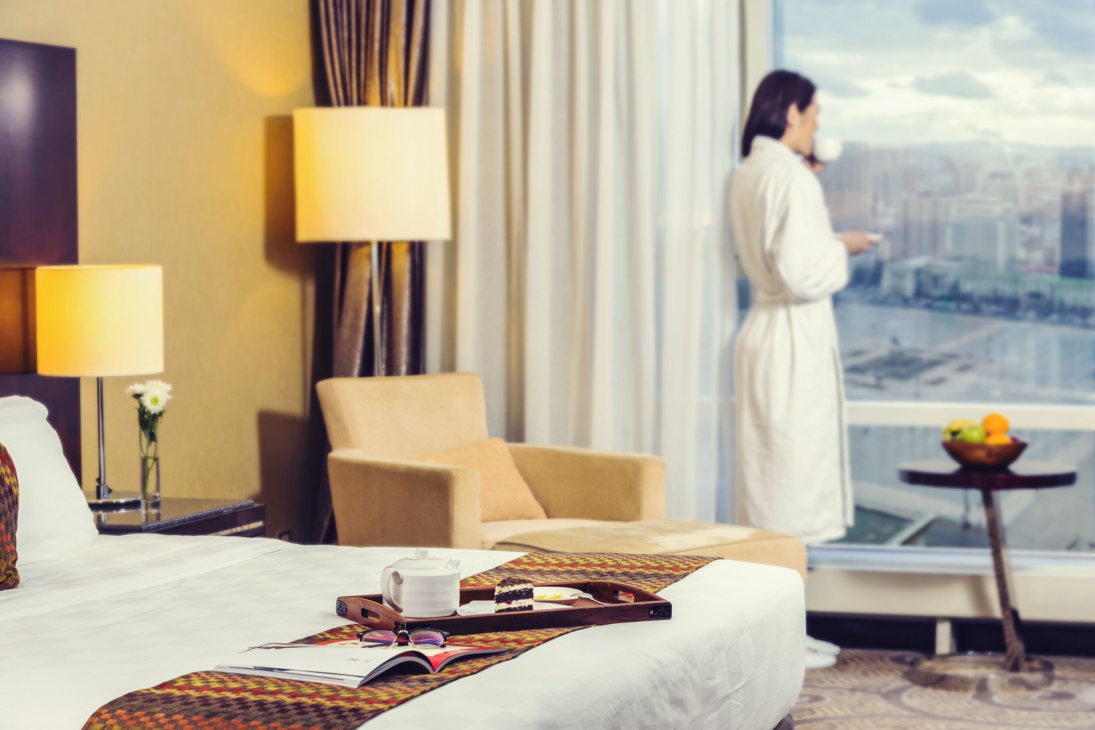 Ein Hotelzimmer mit Bett, Stuhl, kleinem Tisch und Obstschale, mit Blick auf eine Stadt durch ein großes Fenster.