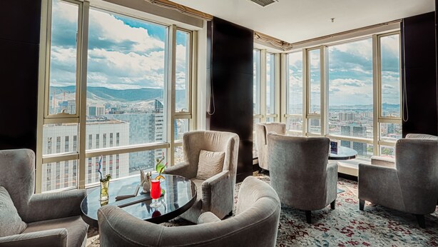 Une chambre d’hôtel moderne dotée de grandes fenêtres donnant sur la ville, de sièges confortables et d’une petite table avec une boisson.