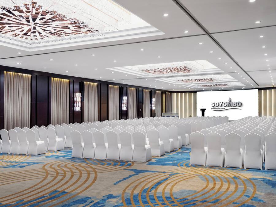 Un salón de conferencias con hileras de sillas blancas, una alfombra estampada y un techo con diseños elaborados.