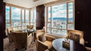 Une chambre d’hôtel située dans un immeuble de grande hauteur, dotée de grandes fenêtres offrant une vue sur la ville, d’une table ronde et de fauteuils confortables.