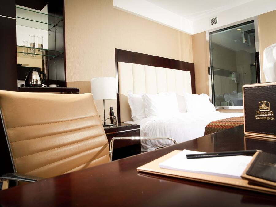 A hotel room with a bed, a desk, a chair, a bedside table, a lamp, a remote control, and a book.
