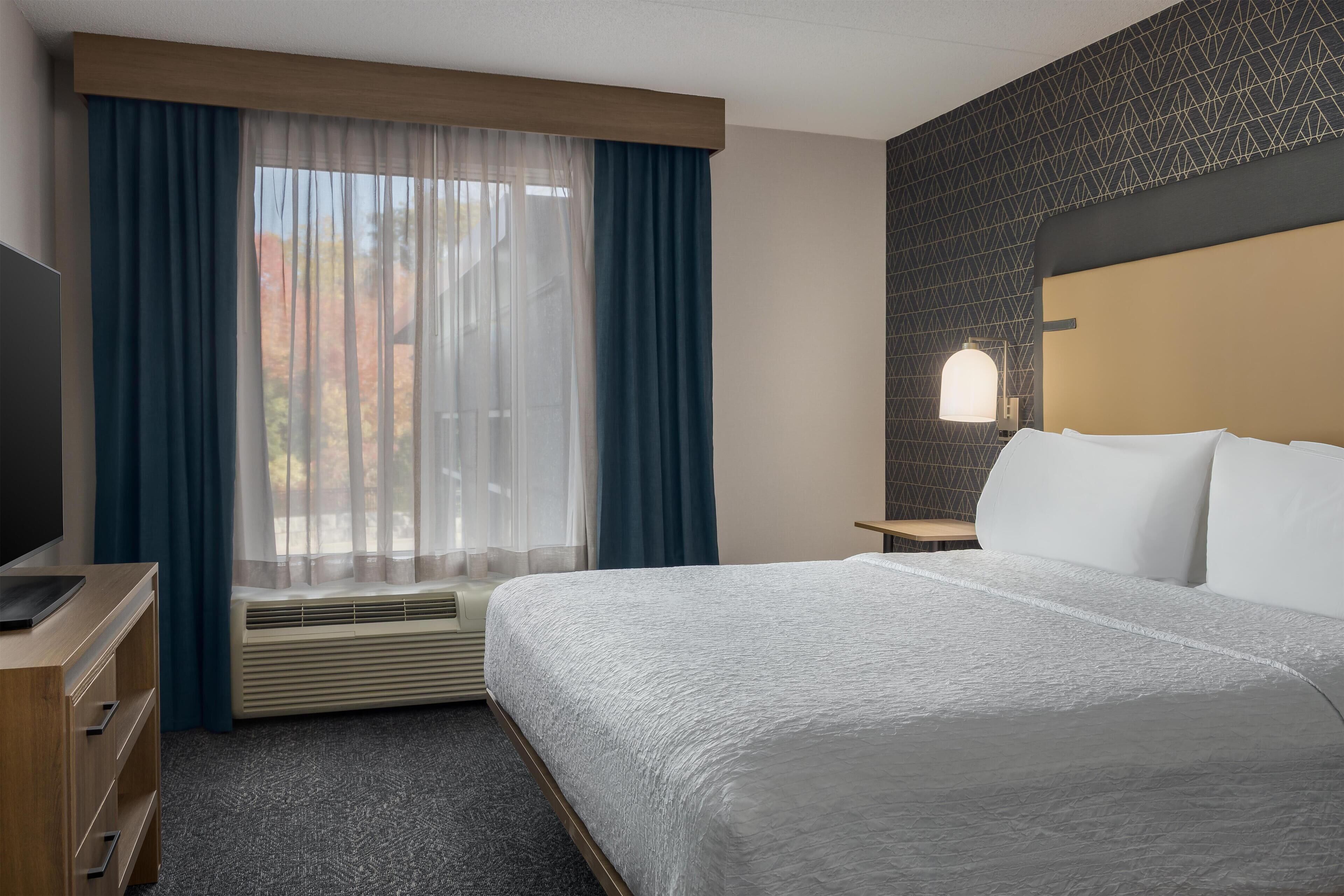 A hotel room with a large bed, a desk with a flat-screen TV, a window with sheer curtains, an air conditioning unit, and a patterned wall.