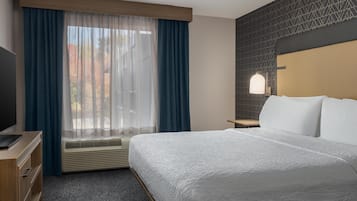 A hotel room with a large bed, a desk with a flat-screen TV, a window with sheer curtains, an air conditioning unit, and a patterned wall.