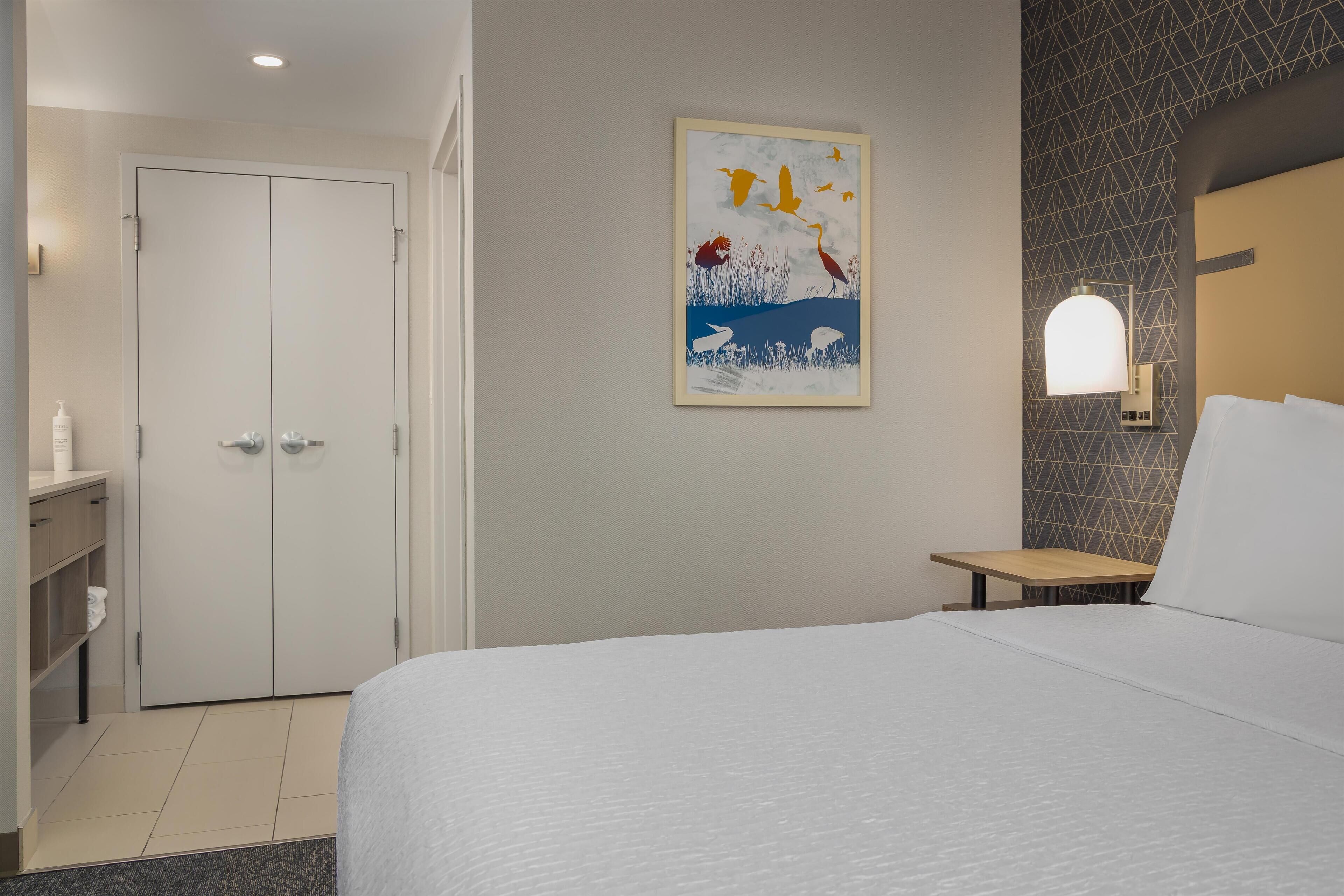 A hotel room with a bed, a desk, a wardrobe, and a wall-mounted light fixture.